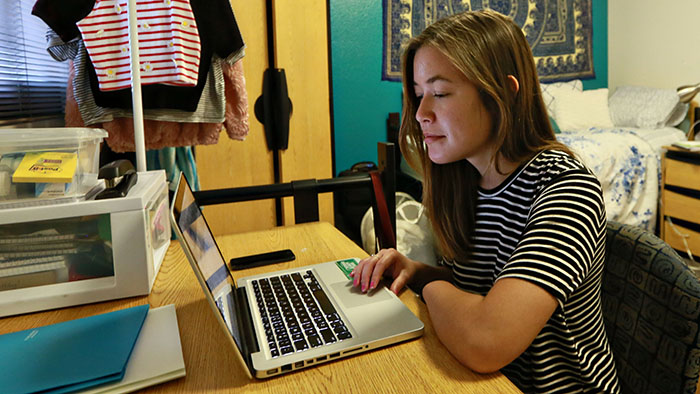 Student focuses on her laptop while studying at a desk in a dorm room setting with clothing and decor in the background.