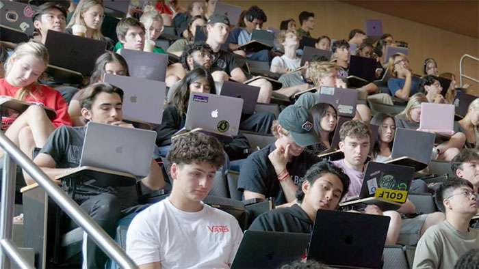 Large group of college students in a lecture hall working on laptops and taking notes during class.