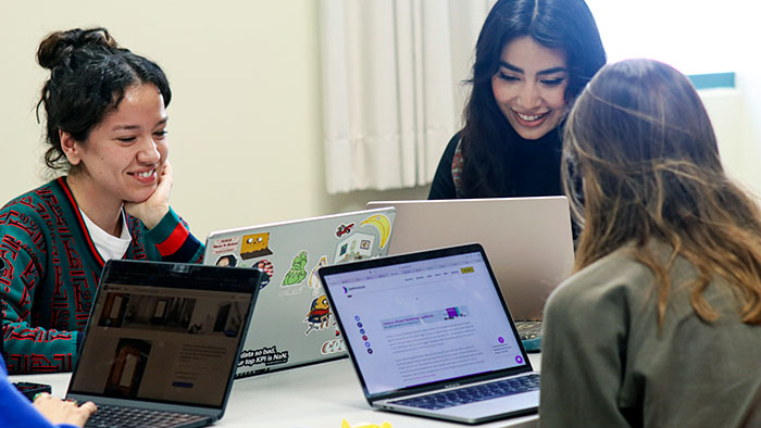 Three students smiling and collaborating at a table with open laptops, actively engaged in group work.