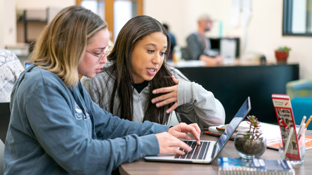 Two students sitting at a round table discussing content on a laptop in a campus study area.